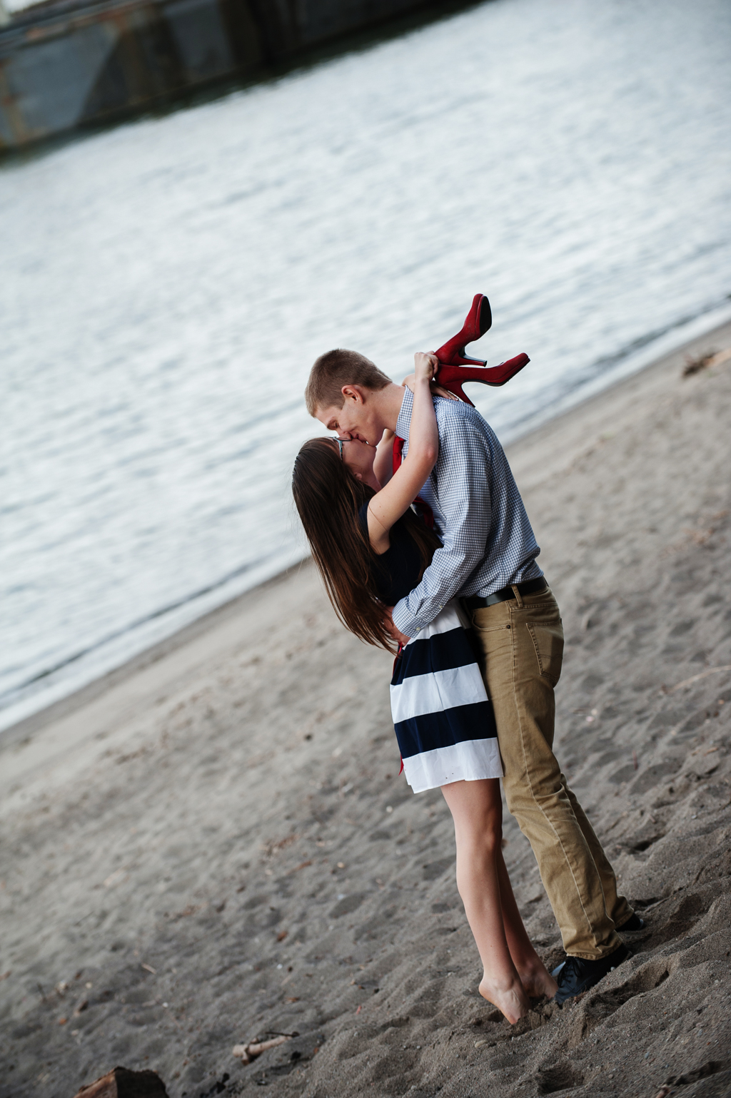 a girl kisses a boy on the beach and she holds her red high heels 