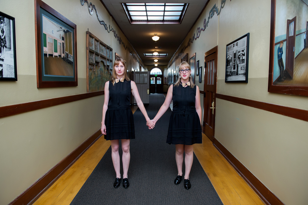 two girls in matching black dresses and bow ties stand like the girls from The Shining in the middle of the hallway at kennedy school