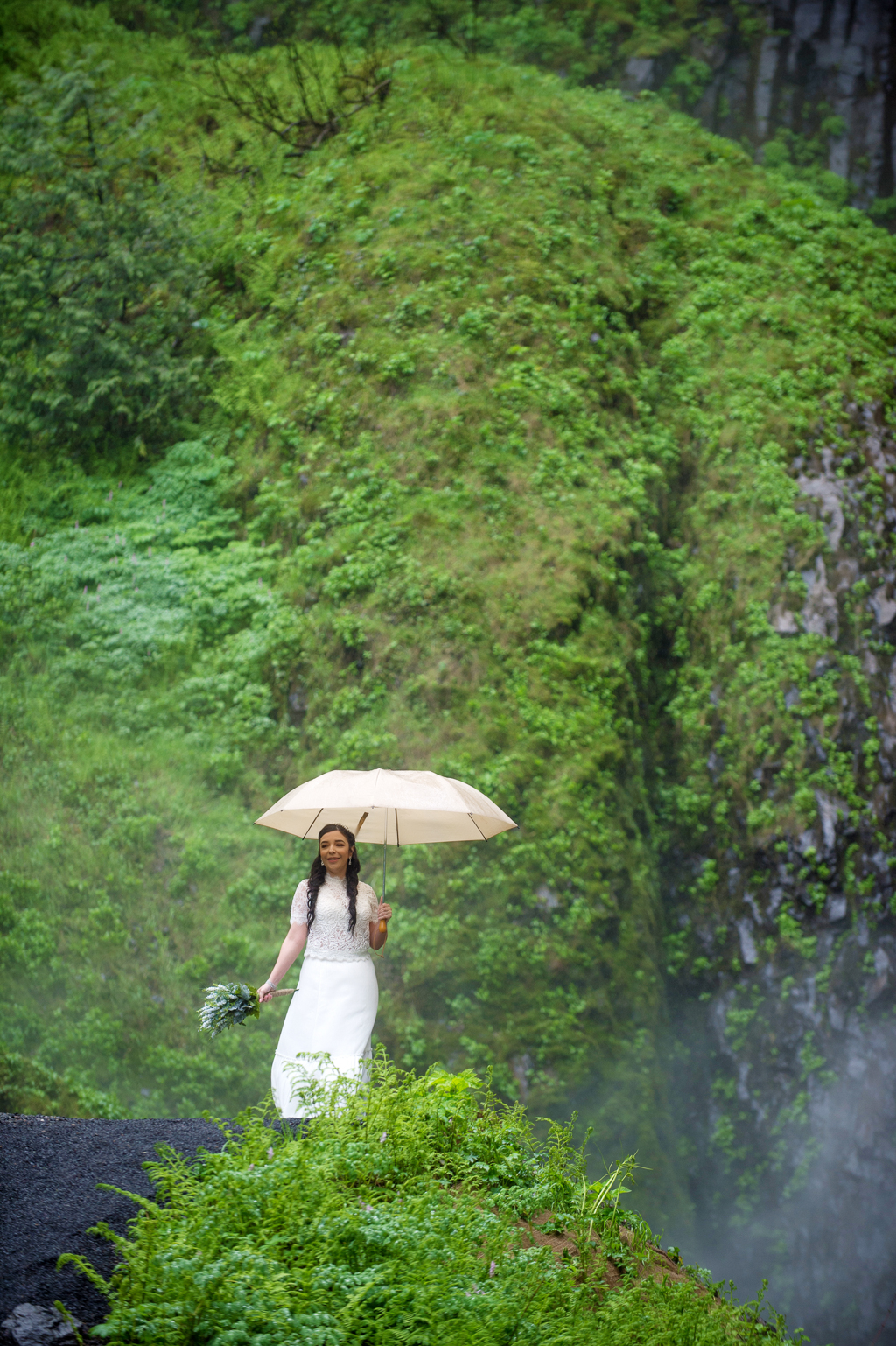 a bride with an umbrella stands on a green moss covered cliff
