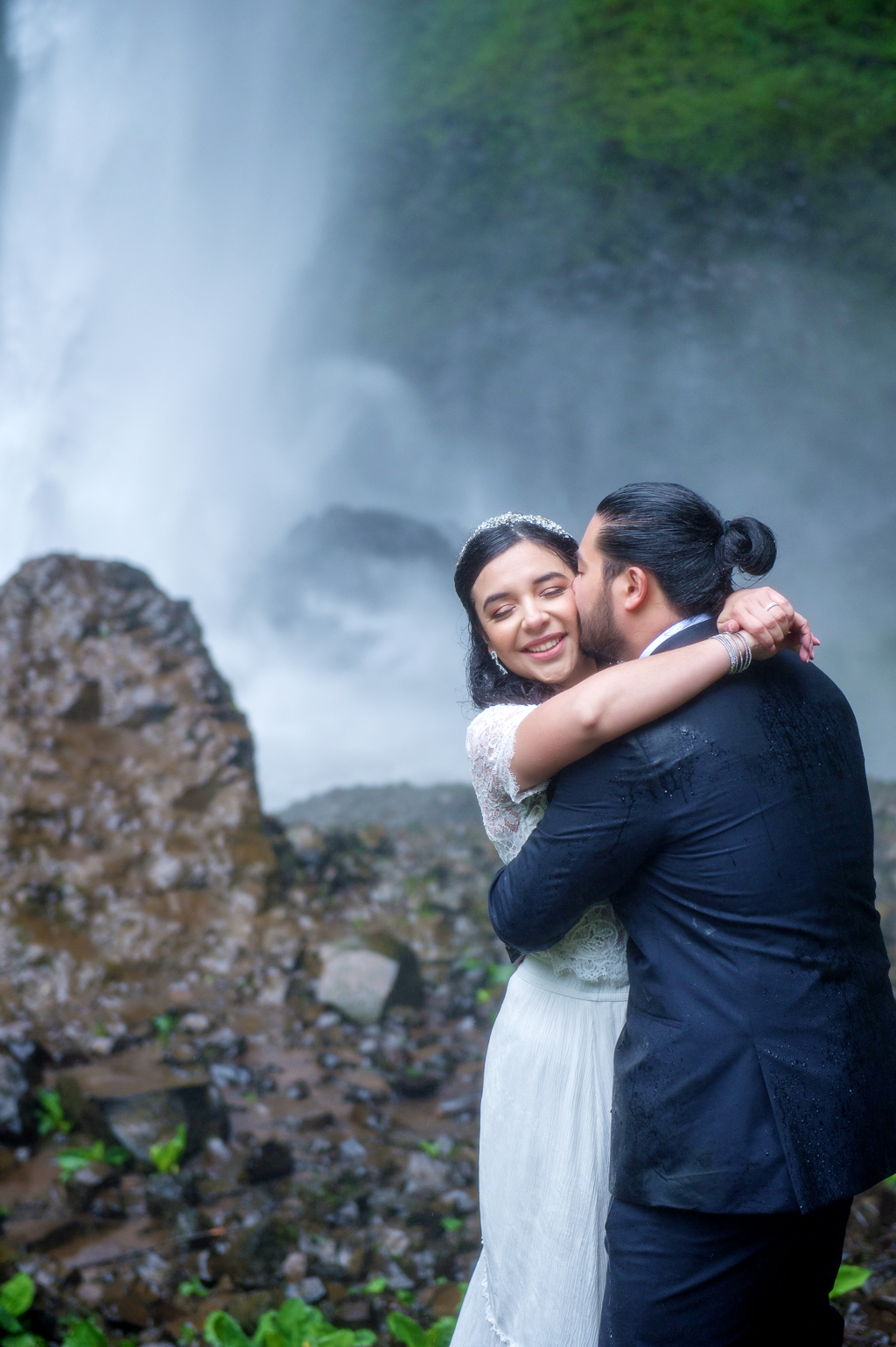 a groom kisses his bride's cheek in front of a waterfall