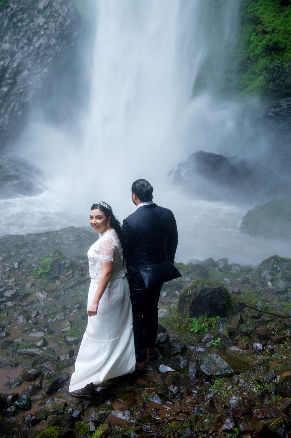 a bride looks over her shoulder as the groom holds her hand and pulls her towards a waterfall