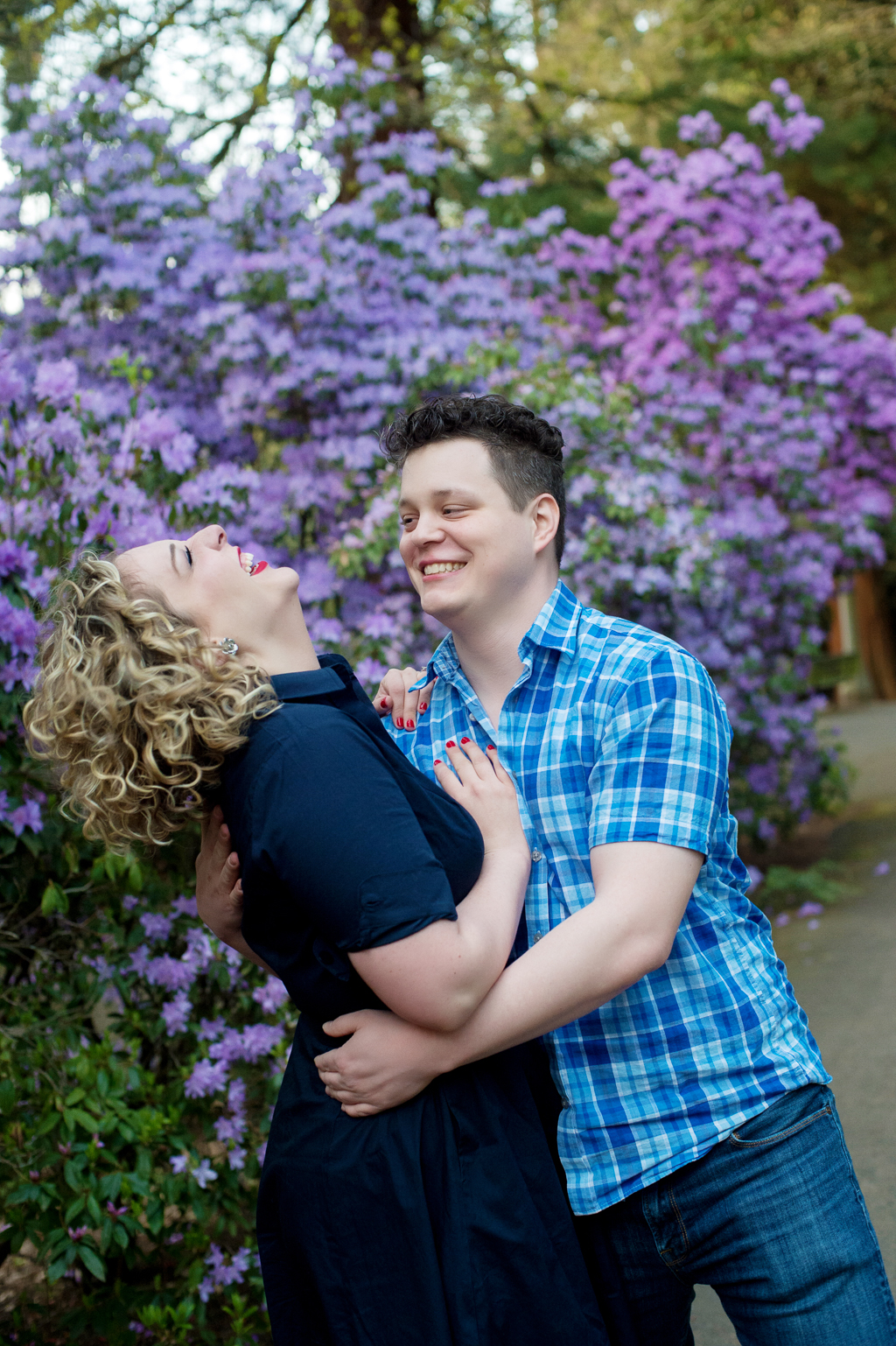 a girl laughs with her head thrown back as her boyfriend hugs her in front of purple flowers
