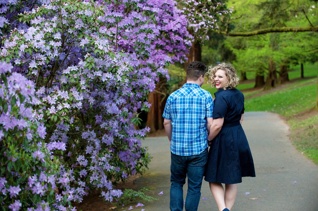 a girl looks over her shoulder while walking with her boyfriend by purple flower bushes at a park