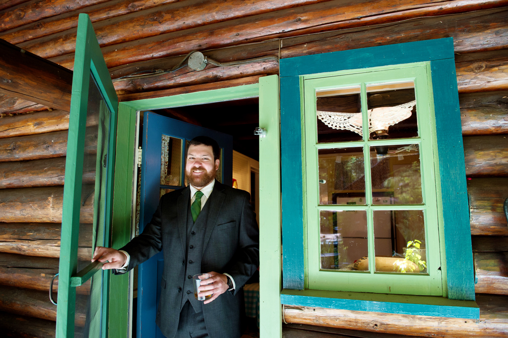 a groom stands in the doorway of a log cabin painted green and turquoise trim