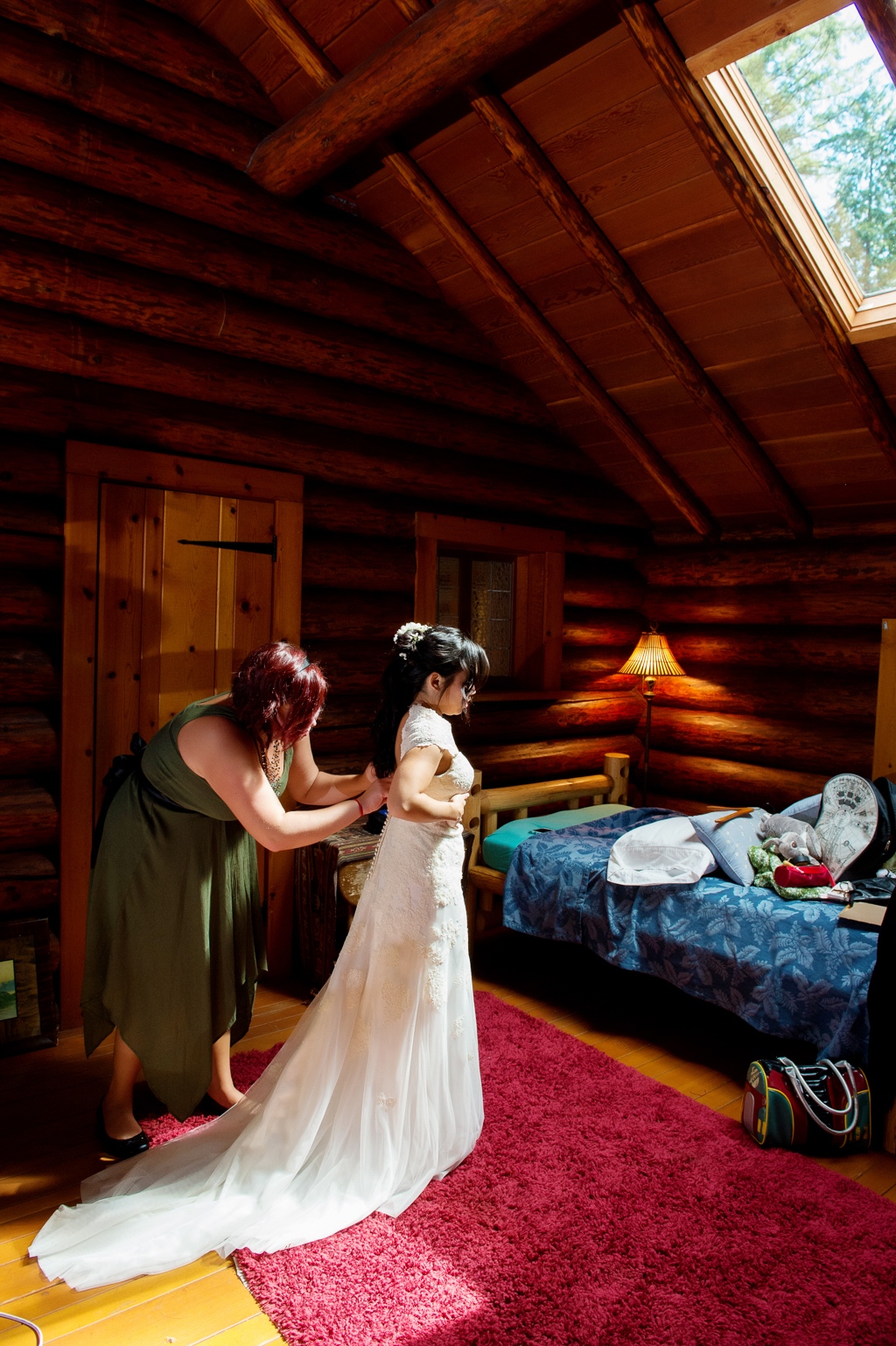 a bridesmaid helps a bride put on her wedding dress in a log cabin