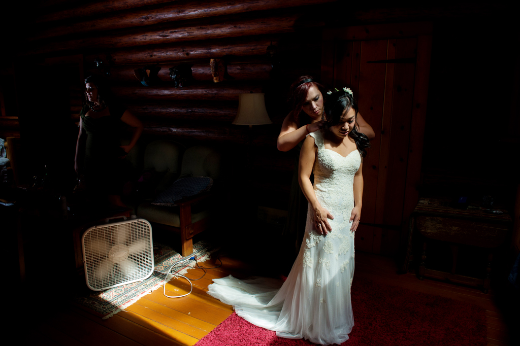 a bridesmaid helps a bride put on her wedding dress in a log cabin