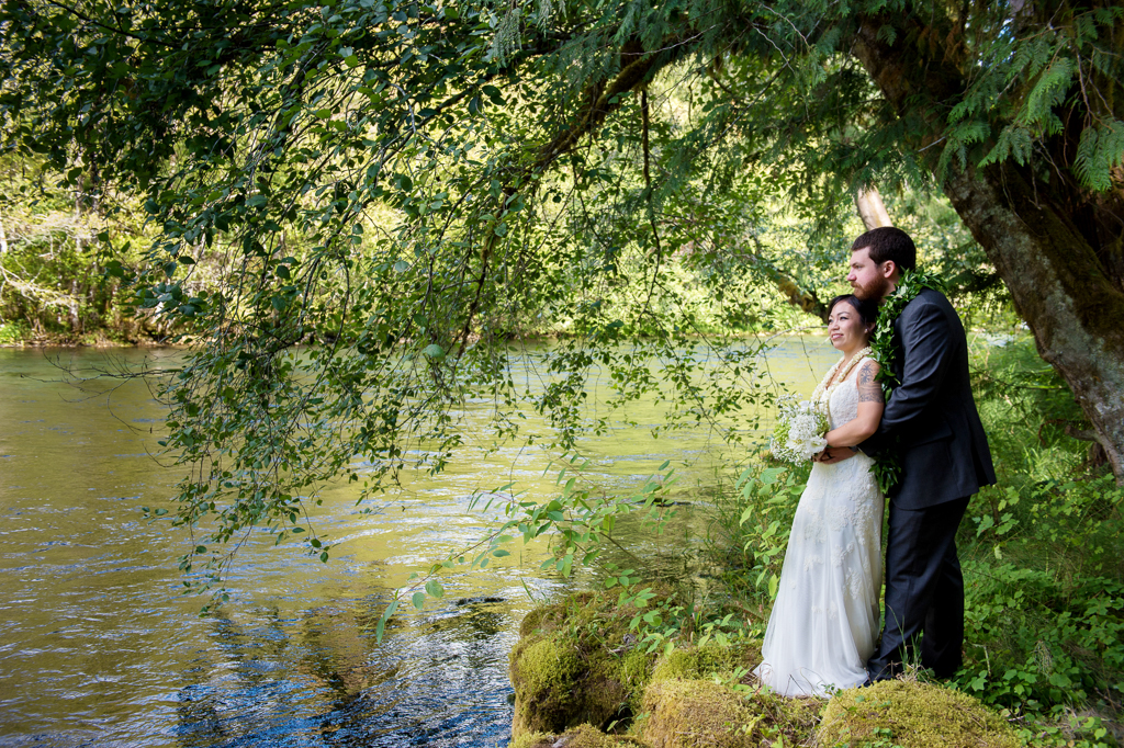 a groom hugs his bride as they stand on the bank next to the mckenzie river