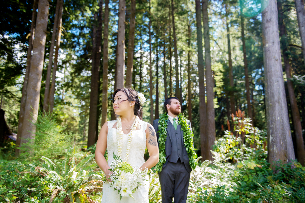 a bride and groom look in opposite directions standing under giant forest trees in the sunshine