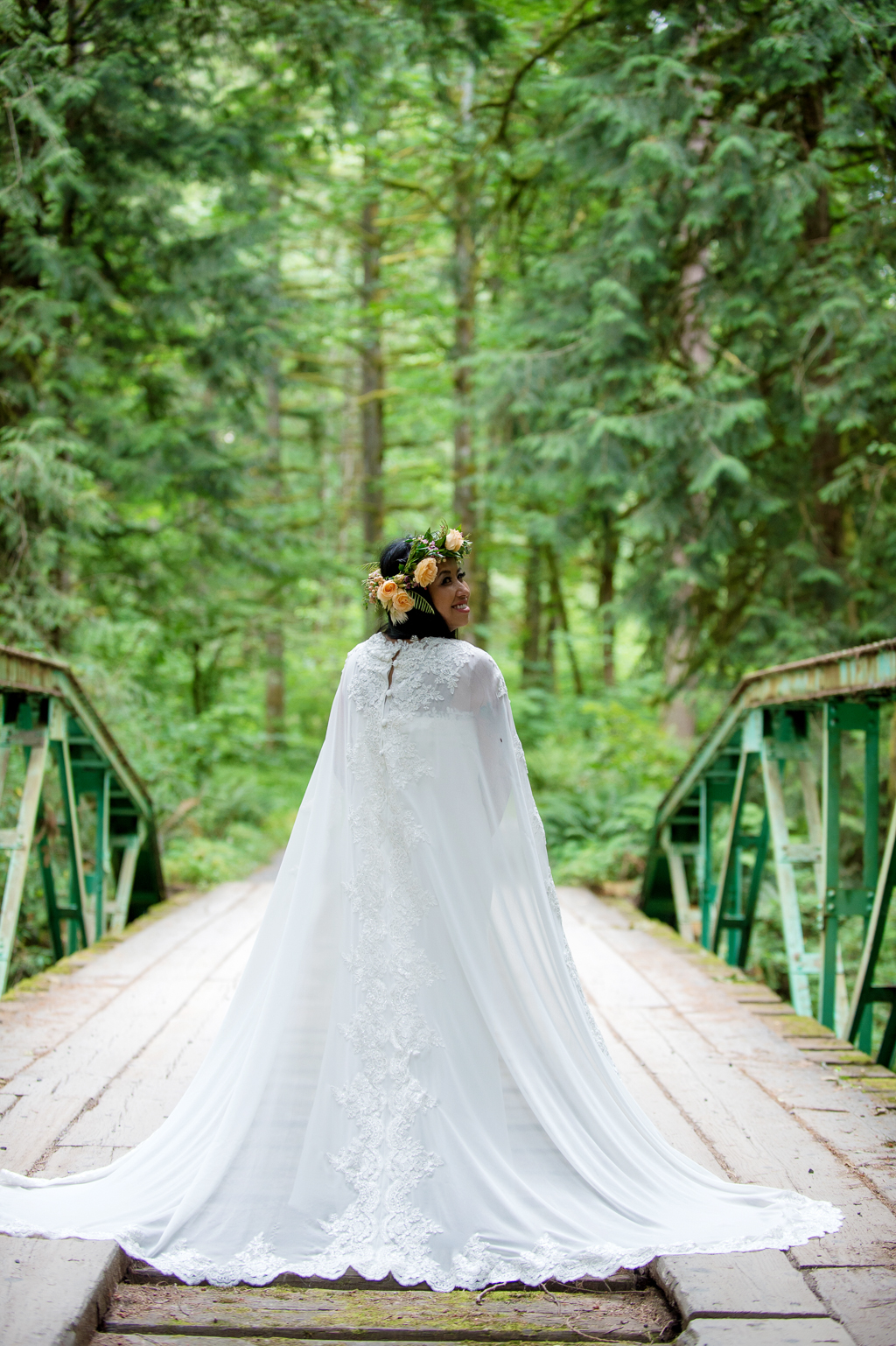 a bride with a very long cape and a flower crown stands on a bridge