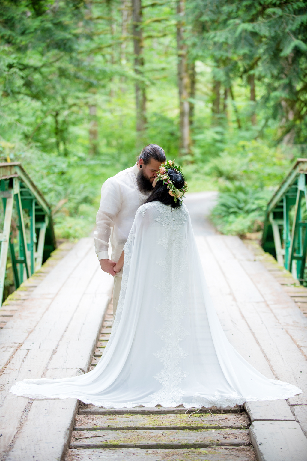 a bride with a very long wedding cape and a flower crown kisses her groom in the middle of a bridge