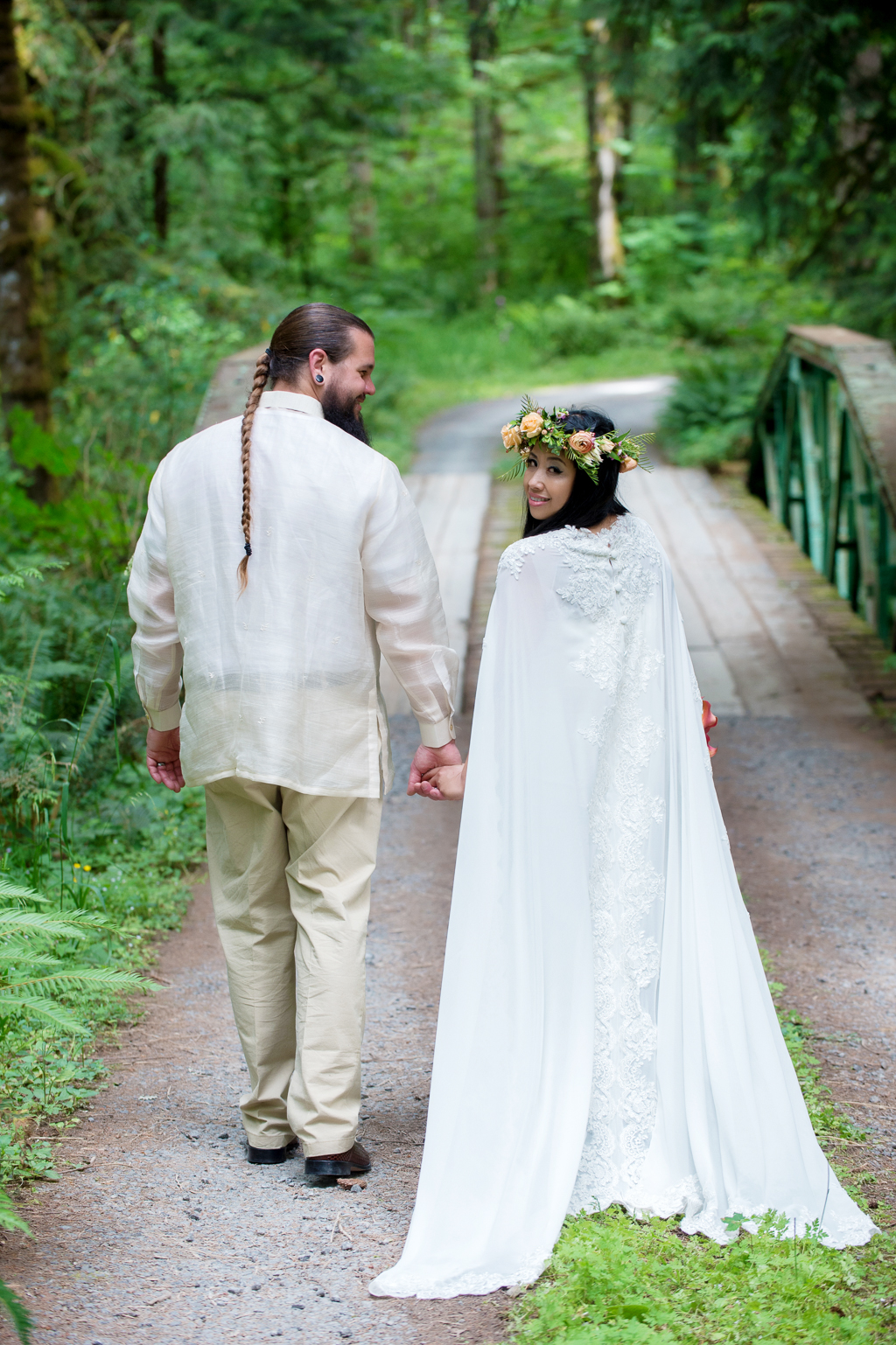 a bride with a very long wedding cape and a flower crown holds hands with her groom who has a very long braid and looks over her shoulder at the camera