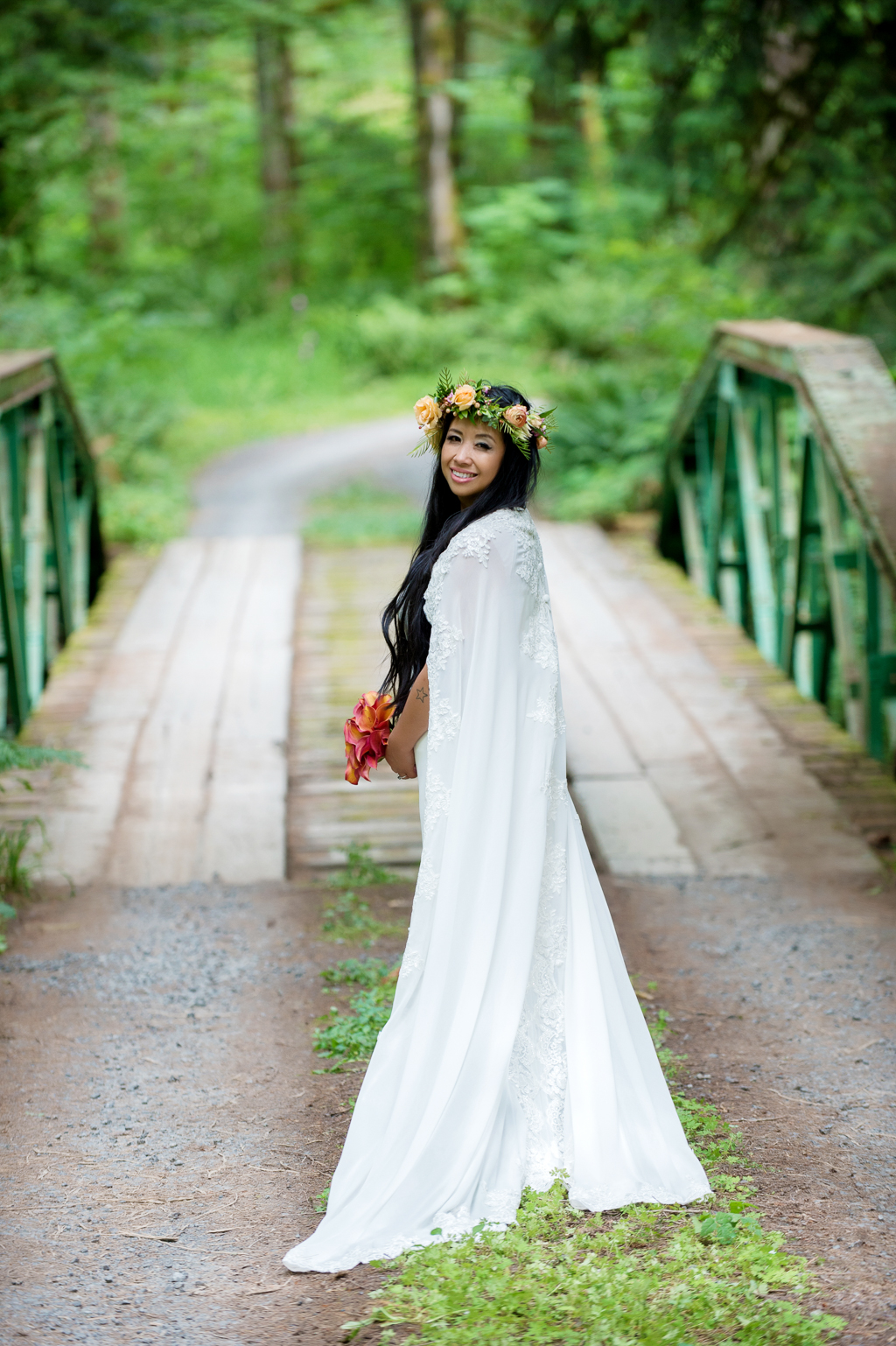 a bride with a very long wedding cape and flower crown stands on a bridge in the woods