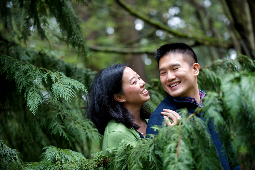 a couple smiles at each other behind evergreen branches