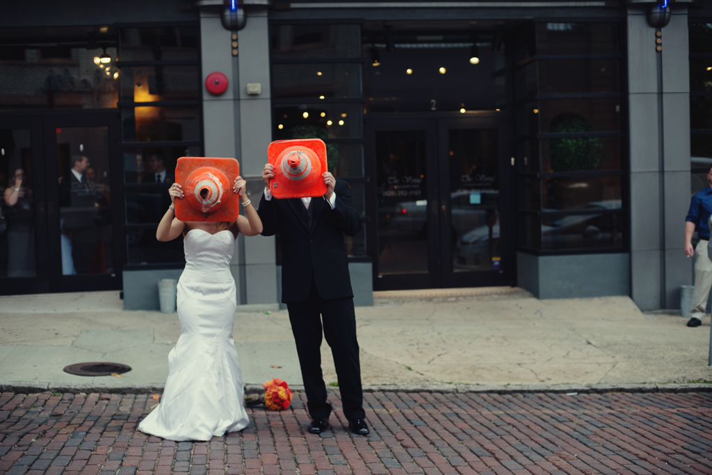 bride and groom cover faces with orange traffic cones