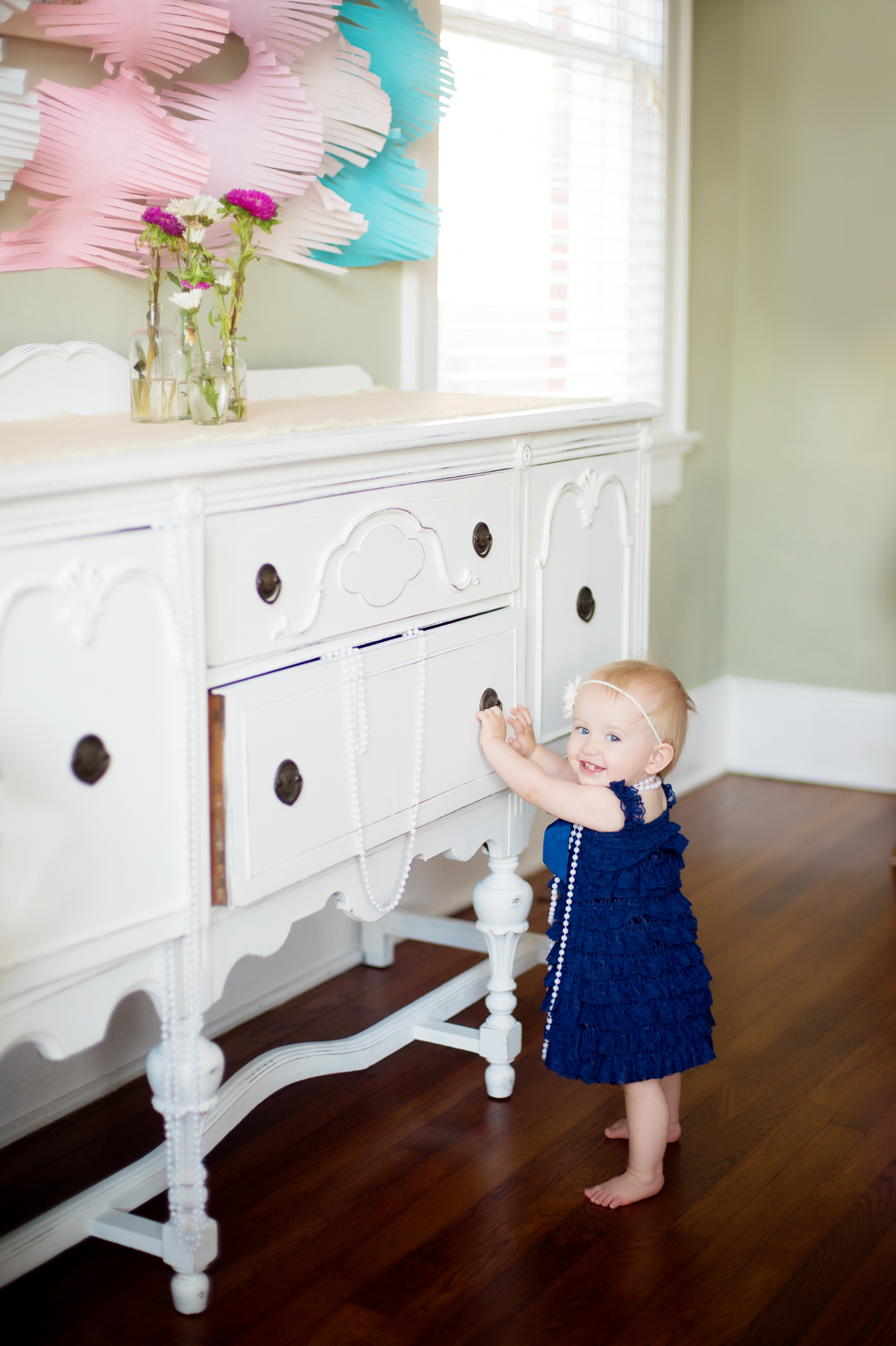 a baby girl in a blue lace romper and pearls stands in front of a white dresser