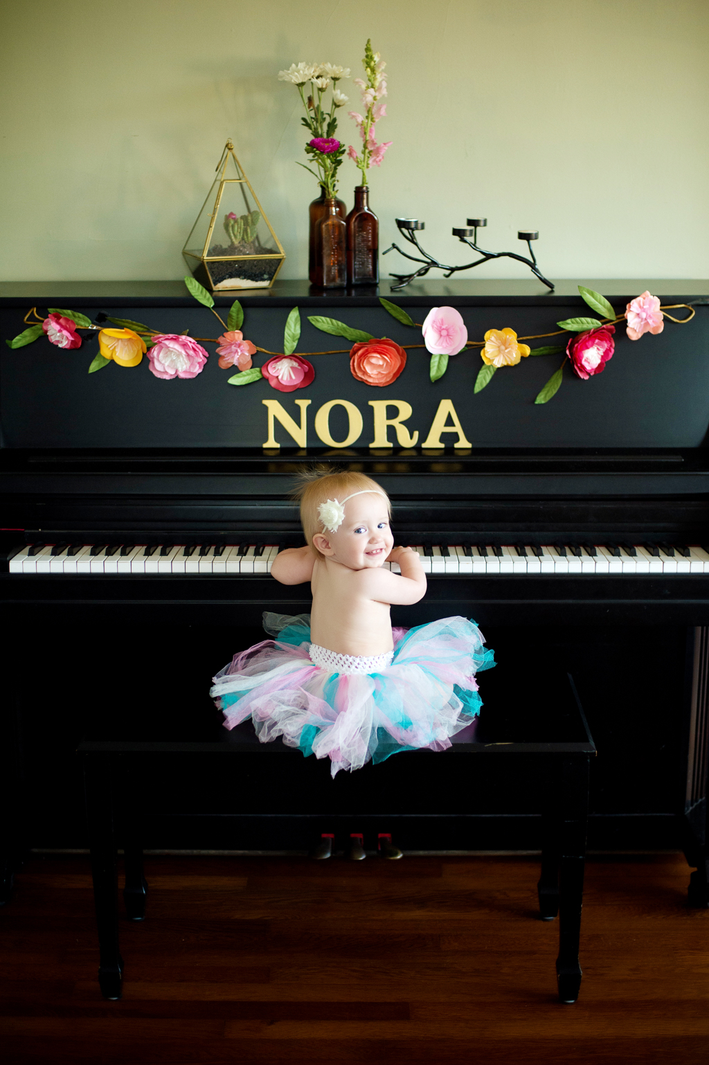 a baby girl in a pink and turquoise tutu looks over her shoulder as she sits at a piano