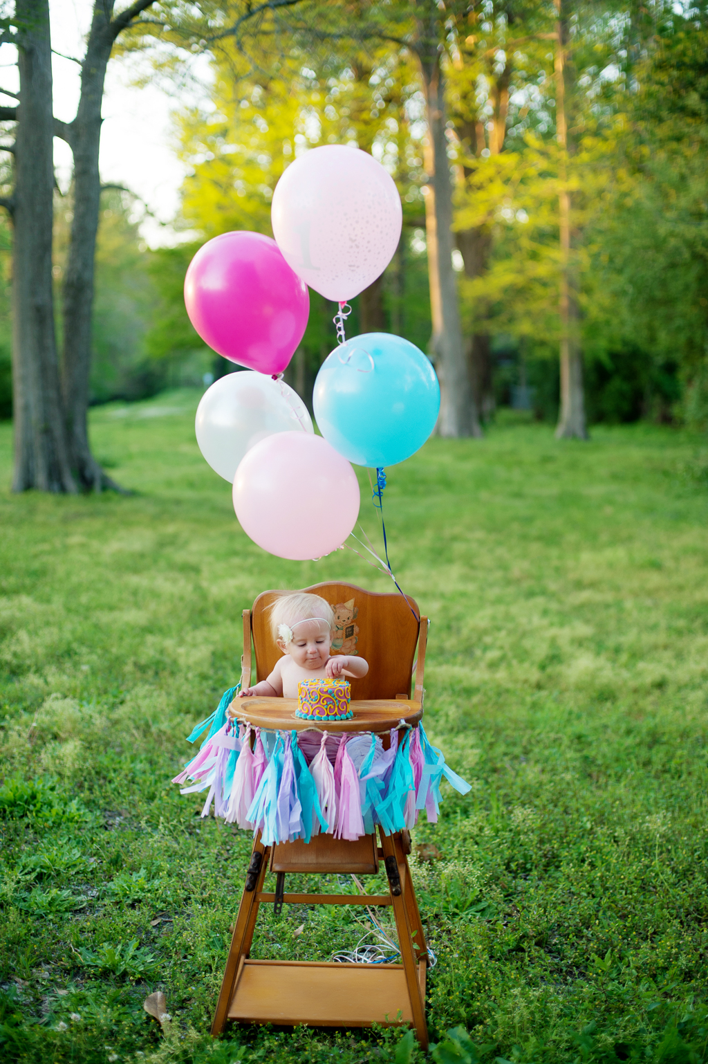 a baby girl sits outside in a high chair covered in balloons and tassels while she eats her birthday cake