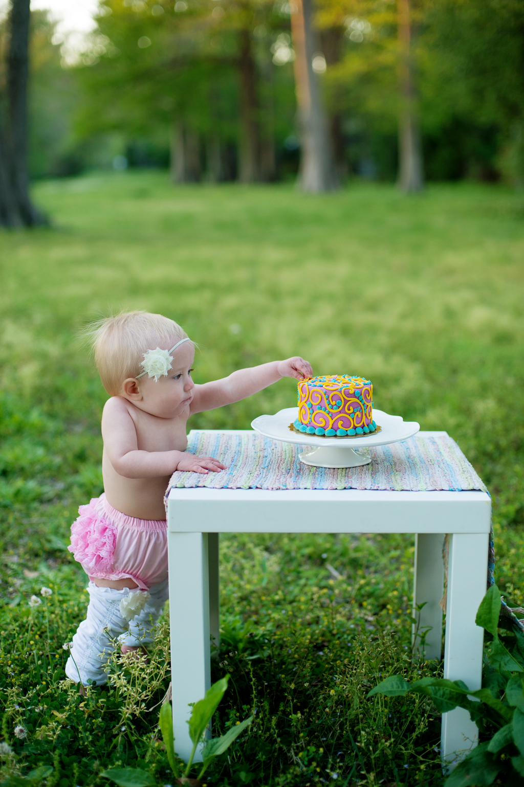 a baby girl in a pink ruffled diaper cover reaches for her confetti birthday cake