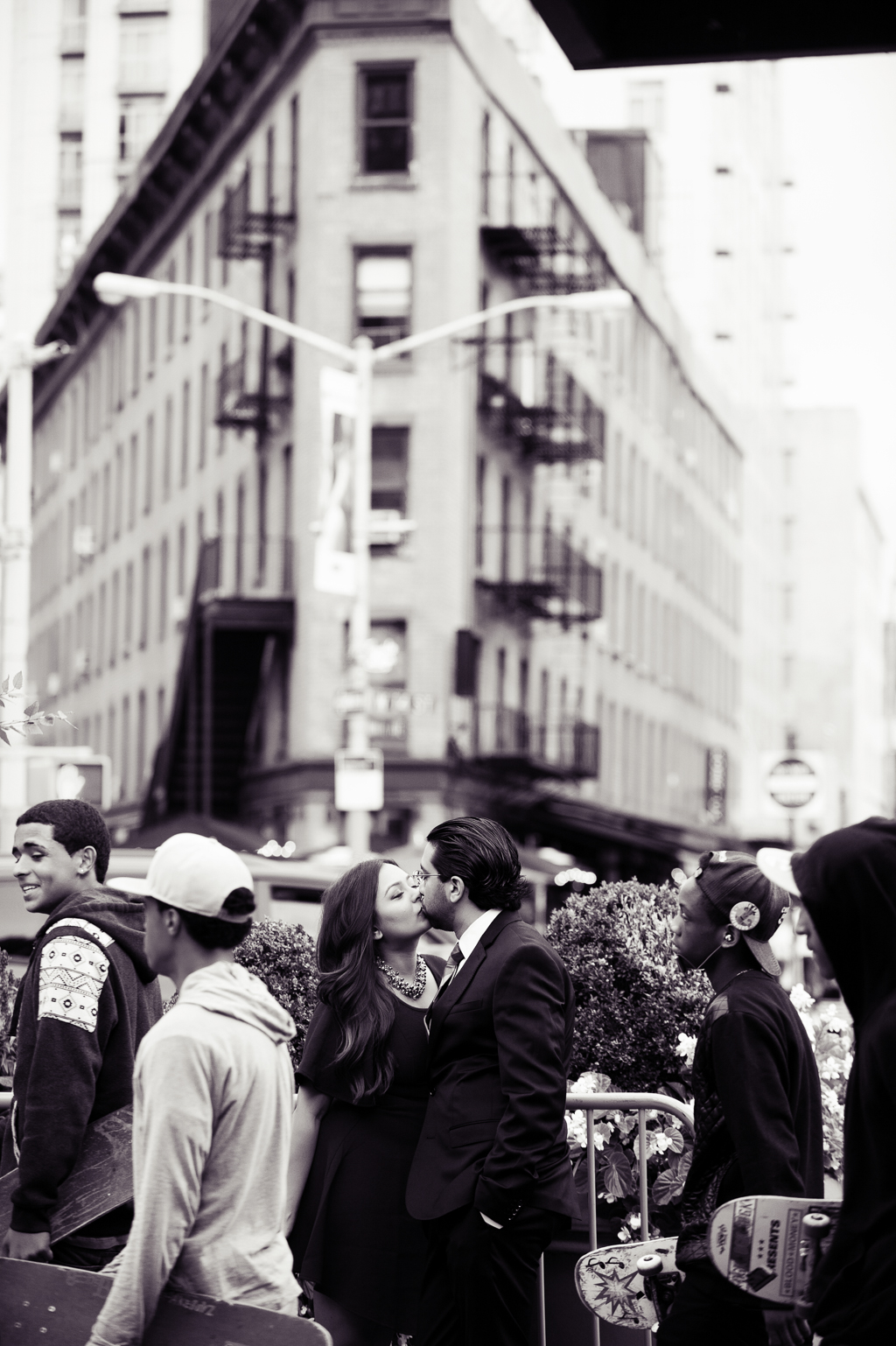 a black and white photo of a couple kissing on a busy nyc street as boys carrying skateboards walk by