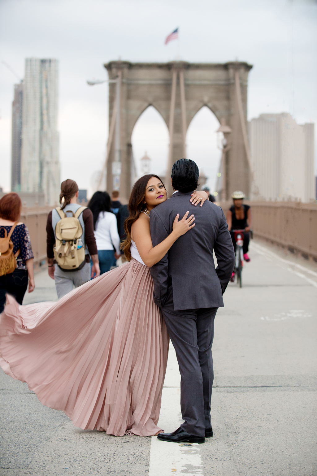 a woman in a long flowy pink skirt hugs around a man in a gray suit on the brooklyn bridge