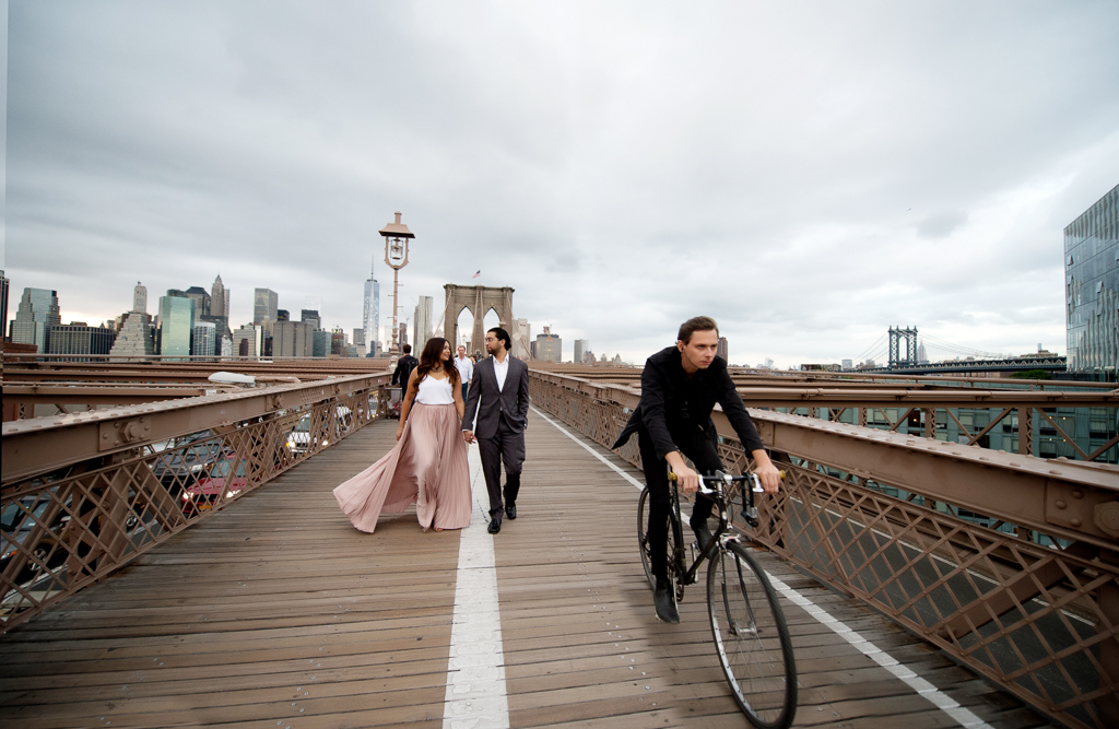 a woman in a long flowy pink skirt walks holding hands on the brooklyn bridge with a man in a gray suit as a cyclist wearing black goes past the frame