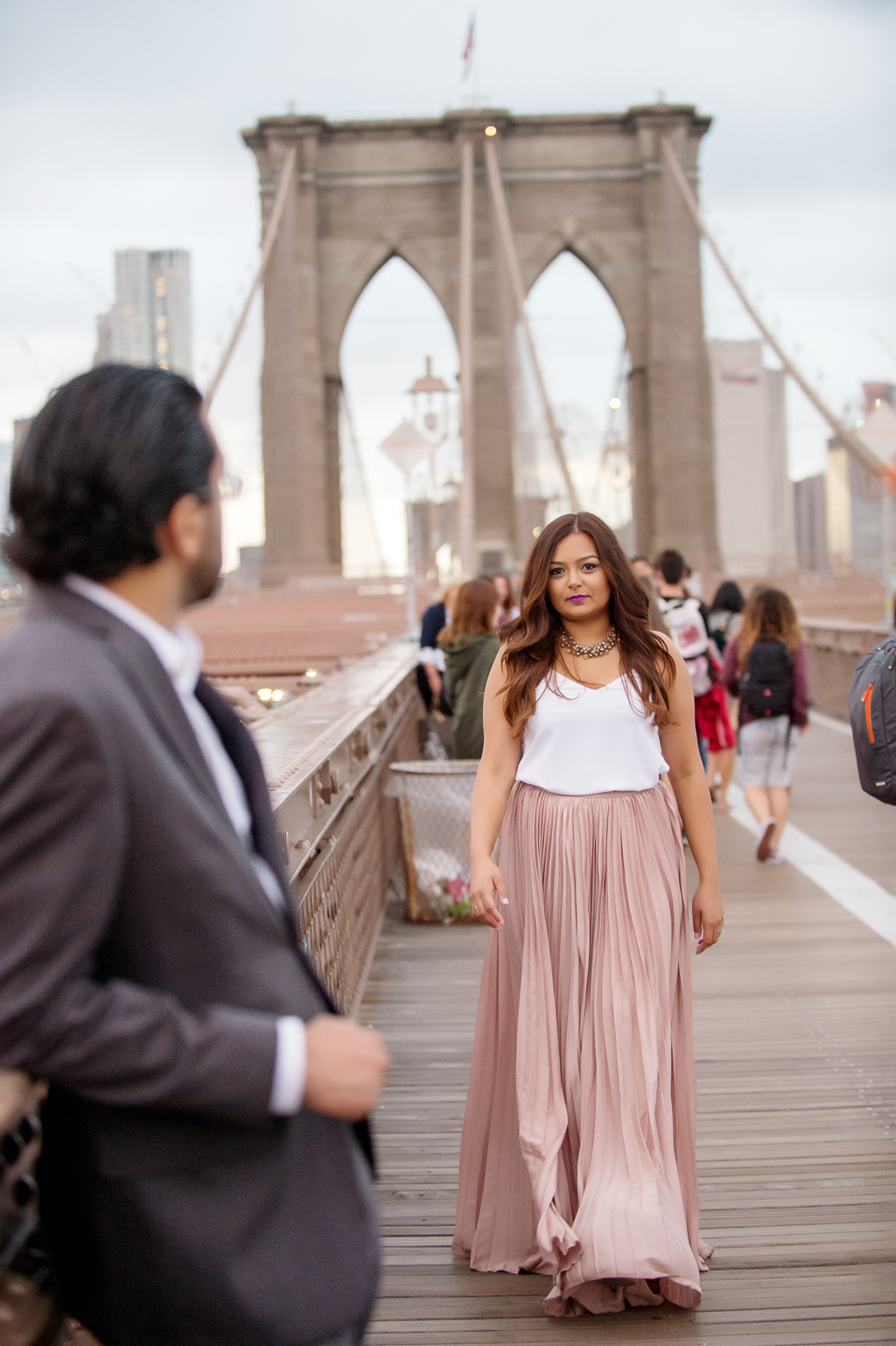 a woman in a flowy pink skirt walks towards a man in a gray suit with the brooklyn bridge in the background