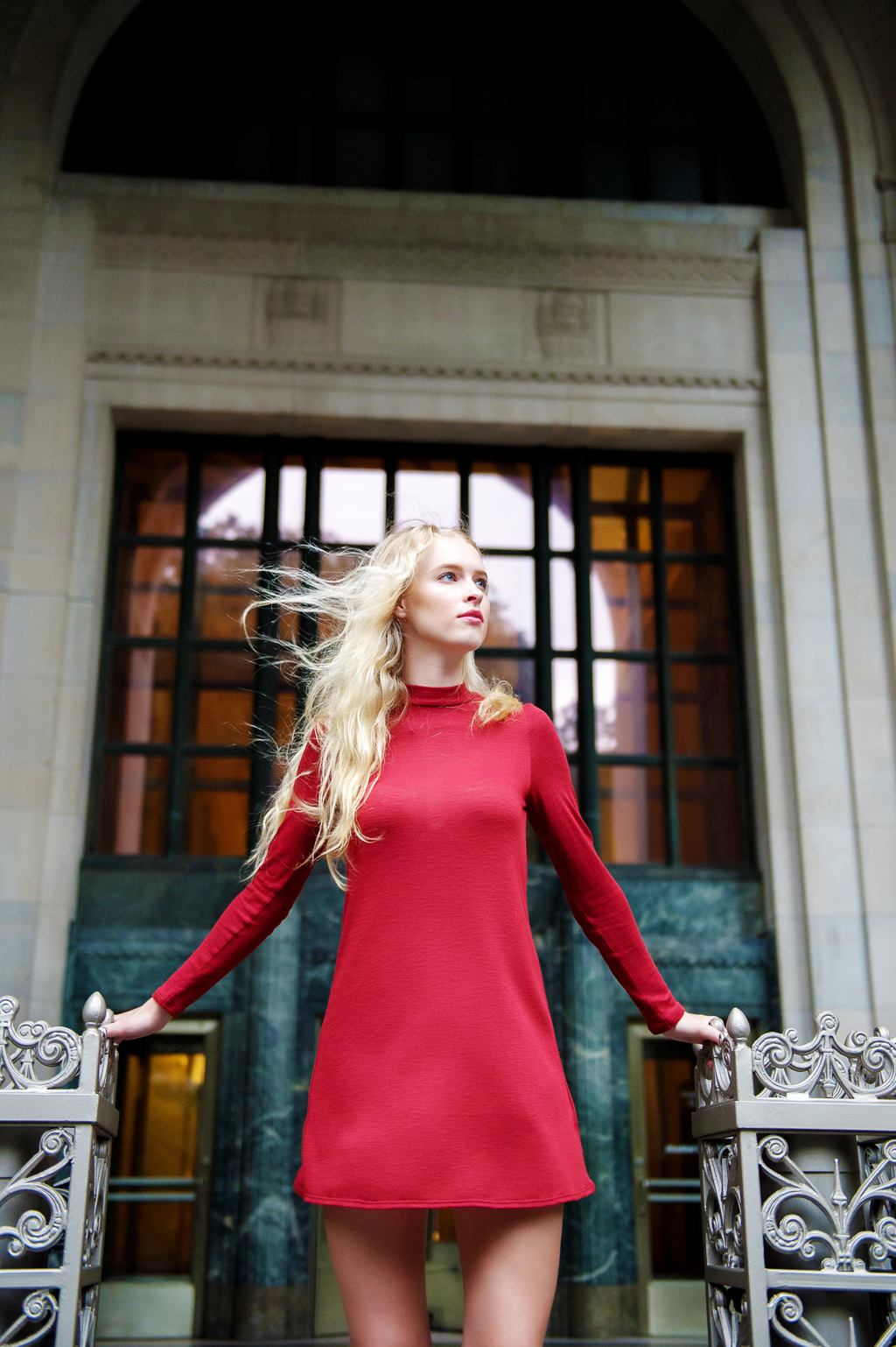 a model in a red dress with long blonde hair stands in front of an old building in NYC