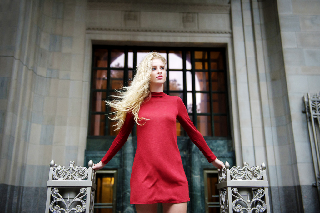 a model in a red dress with long blonde hair stands in front of an old building in NYC