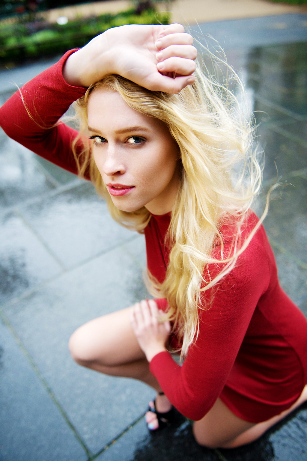a model in a red dress and long blonde hair squats on a rain covered sidewalk