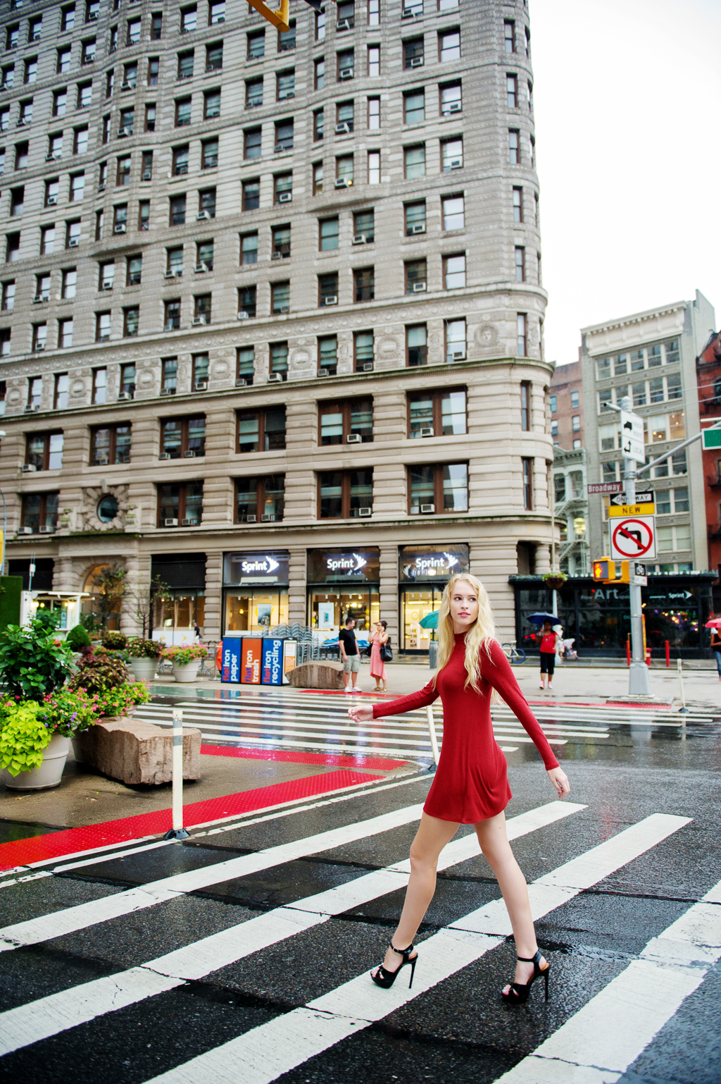 a model in a red dress and long blonde hair crosses the street in front of the flat iron building