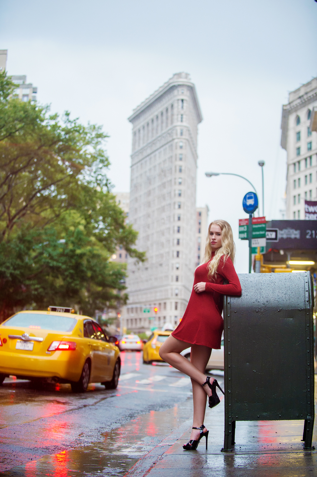 a model in a red dress with long blonde hair leans against a post office mailbox in front of the flat iron building in nyc as yellow taxis drive by