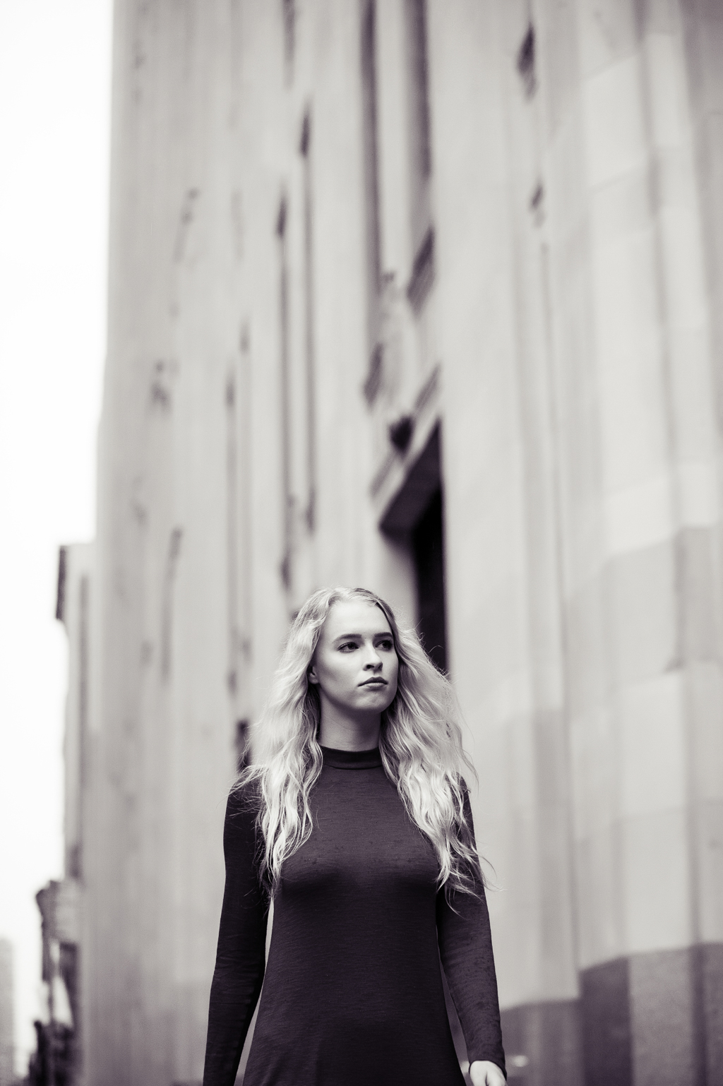 a model with long blonde hair walks down a sidewalk in nyc with a tall building towering above