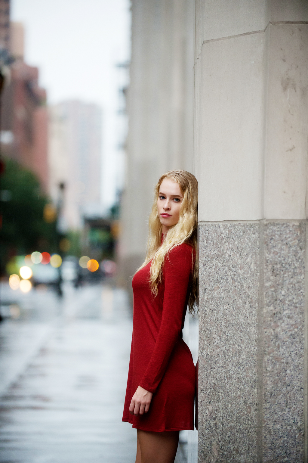 a model in a red dress with long blonde hair leans against a building in nyc