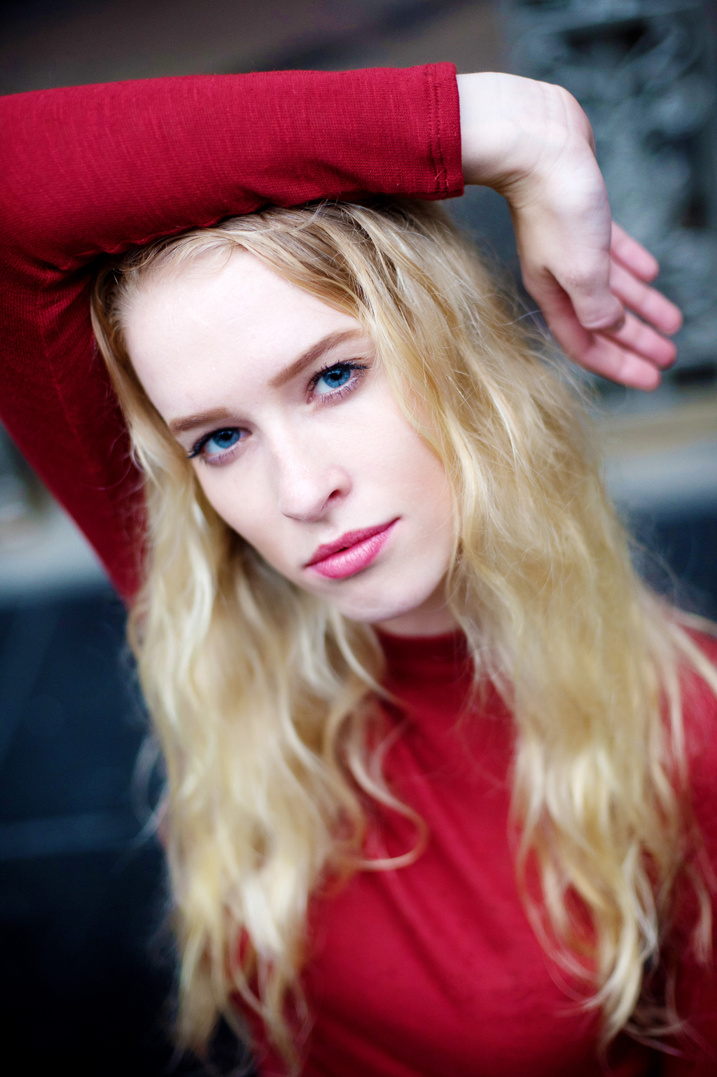 a model in a red dress with long blonde hair looks intently at the camera with her arm over her head