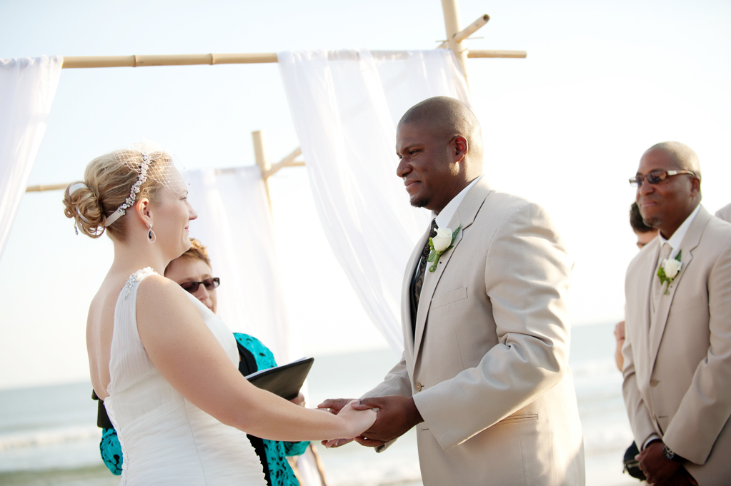a bride and groom stand holding hands at their wedding ceremony on a beach in front of a wooden arbor draped with white cloth