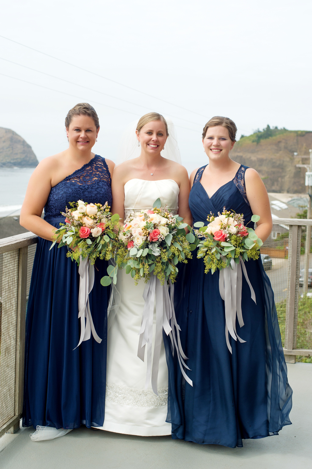 bride stands between 2 bridesmaids in navy blue dresses holding green pink and ivory bouquets