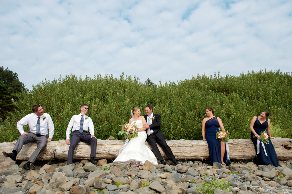 bridesmaids and groomsmen in navy and gray sit on a fallen log on the beach