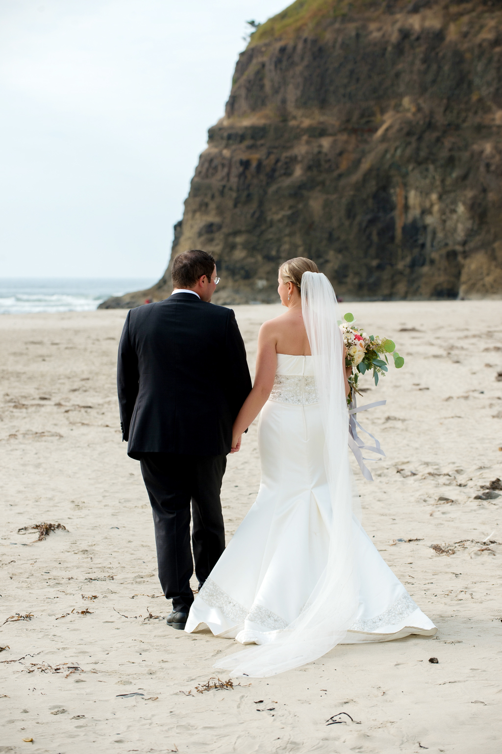 bride with a very long veil holds hands with the groom as they walk towards the ocean along the beach