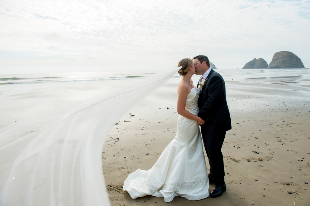 a bride and groom kiss on the beach as her very long veil flies in the wind towards the camera