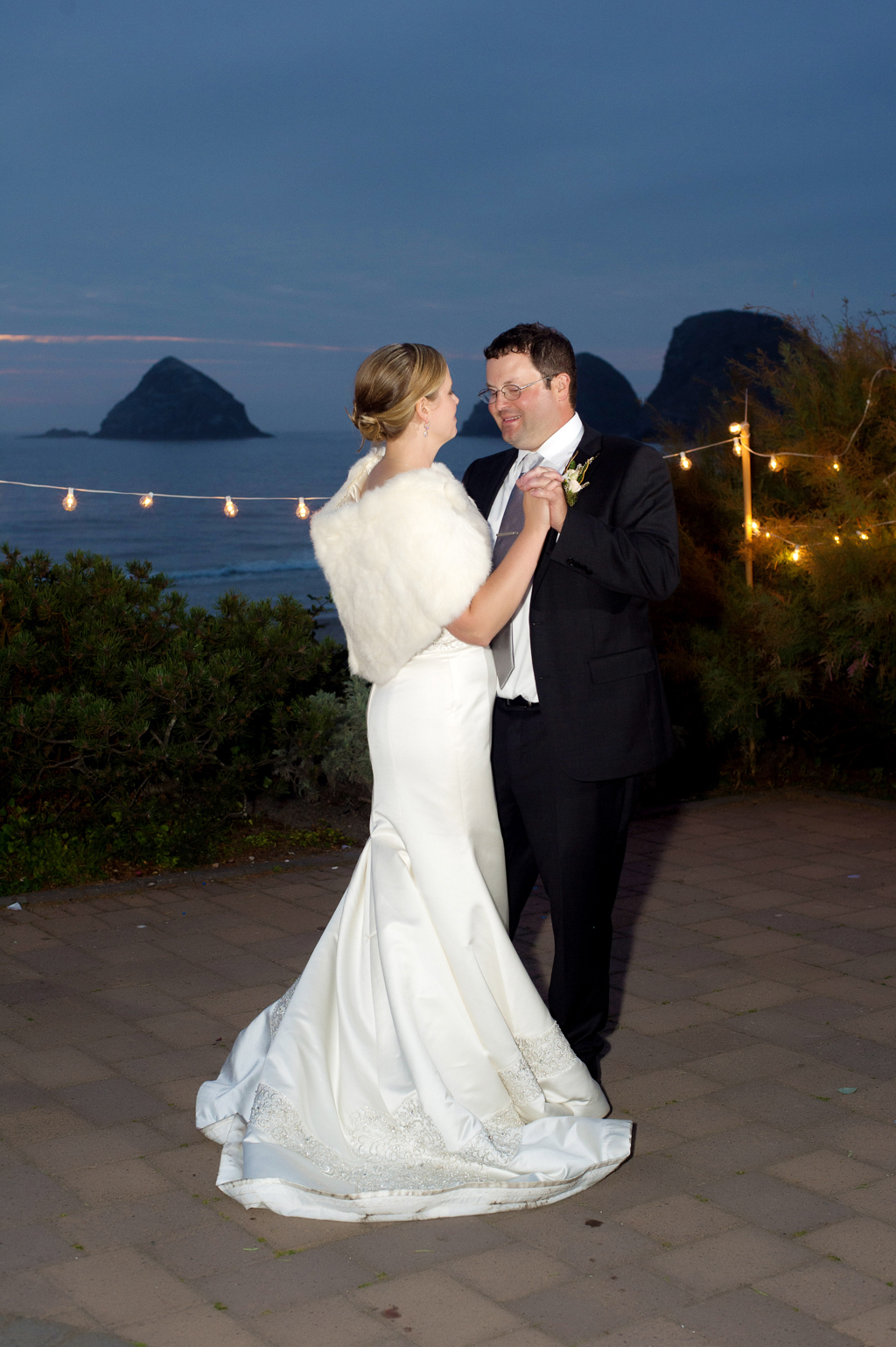 a bride wearing a white fur shawl and groom have their first dance surrounded by cafe lights with the pacific ocean in the background