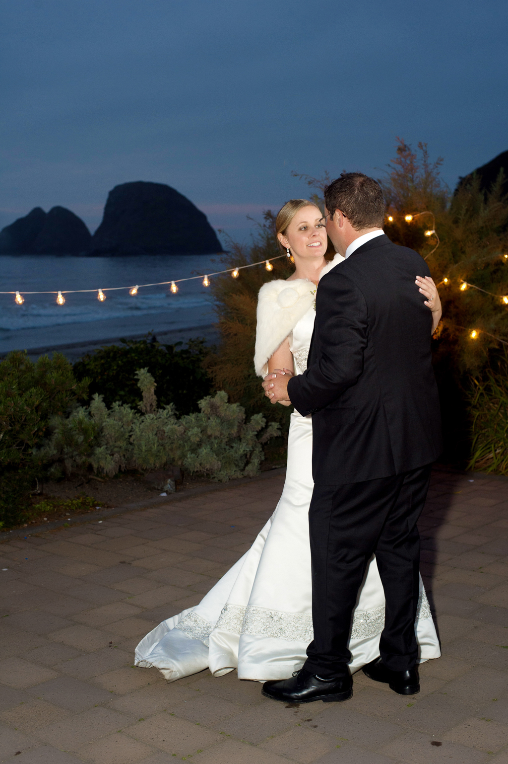 a bride wearing a white fur shawl and groom have their first dance surrounded by cafe lights with the pacific ocean in the background