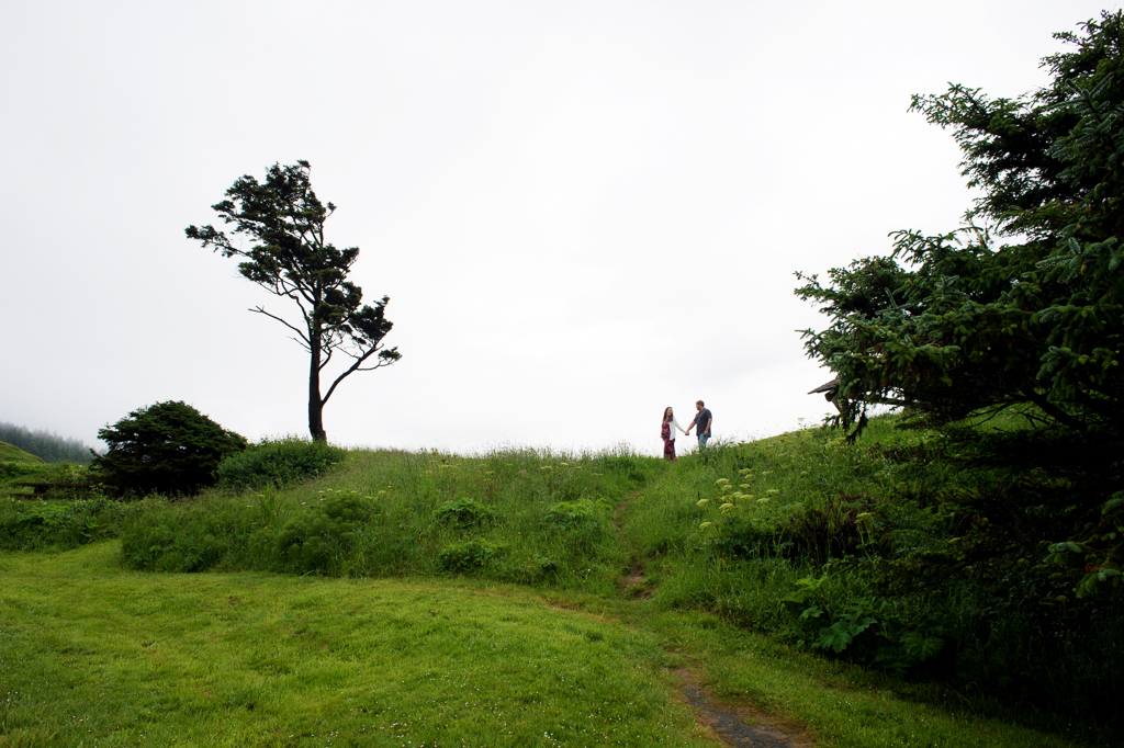 a man and a pregnant woman hold hands and walk across the horizon towards a wind swept tree at ecola state park