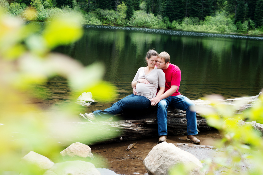 a man and a pregnant woman hug together sitting on a fallen log at mirror lake