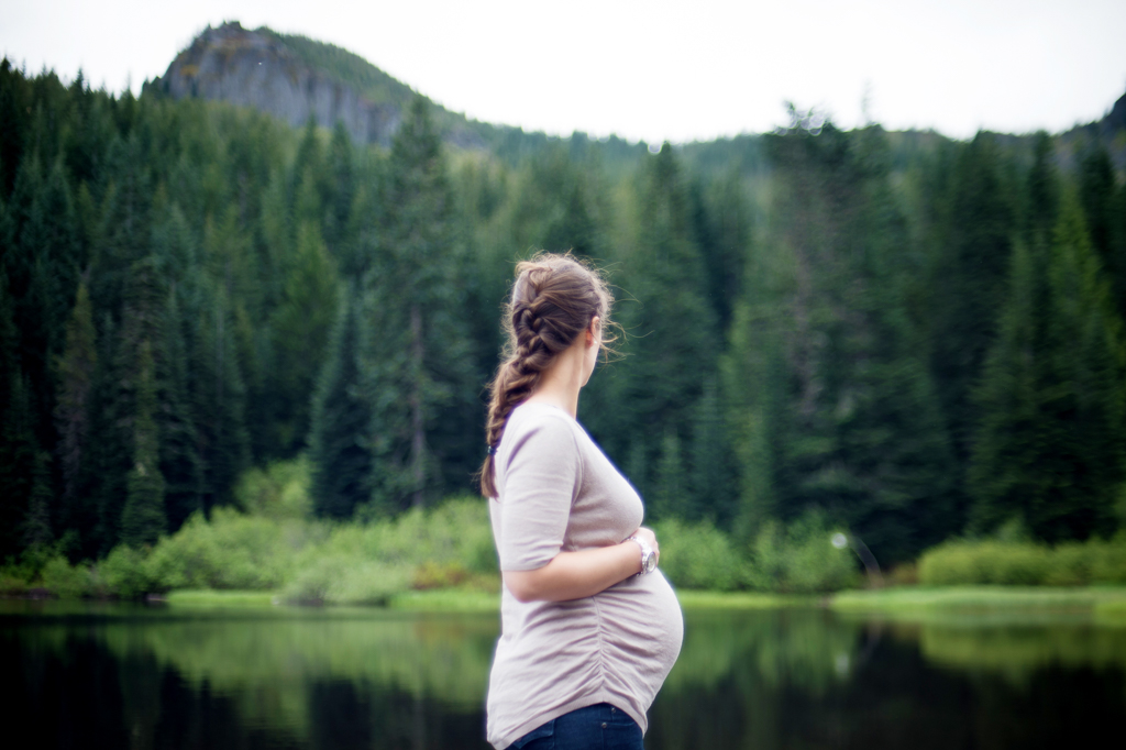 a pregnant woman with a pretty french braid holds her belly and looks out over mirror lake