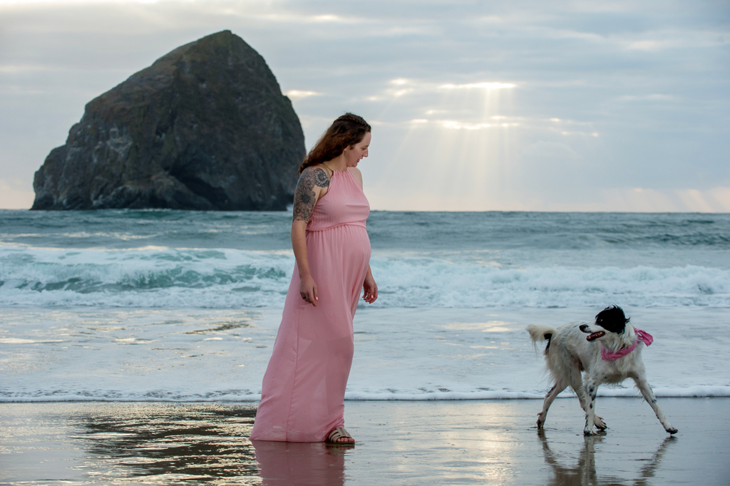 A pregnant woman in a flowy pink dress walks through the ocean's edge following her dog