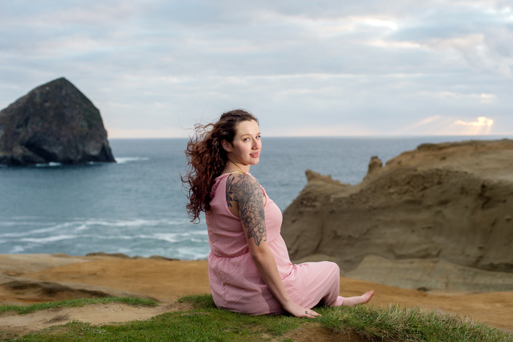 a pregnant woman in a pink dress sits on the edge of the dune that overlooks the ocean