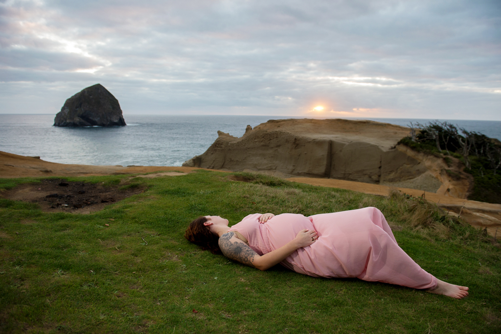 an expecting mother lays in the grass on top of the dune watching the sunset over the ocean