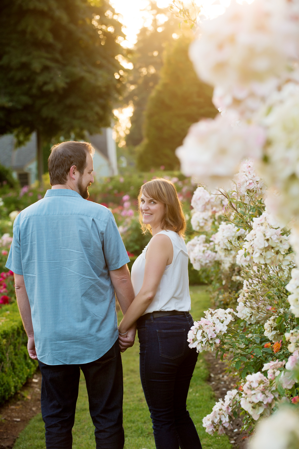 a woman looks over her shoulder at the camera while her boyfriend holds her hand and looks at her surrounded by white roses