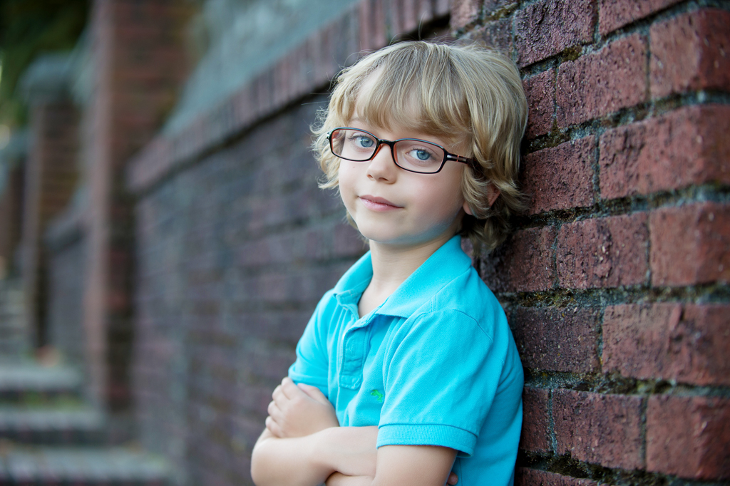 a little boy in glasses wearing a blue shirt with blue eyes leans against a brick wall