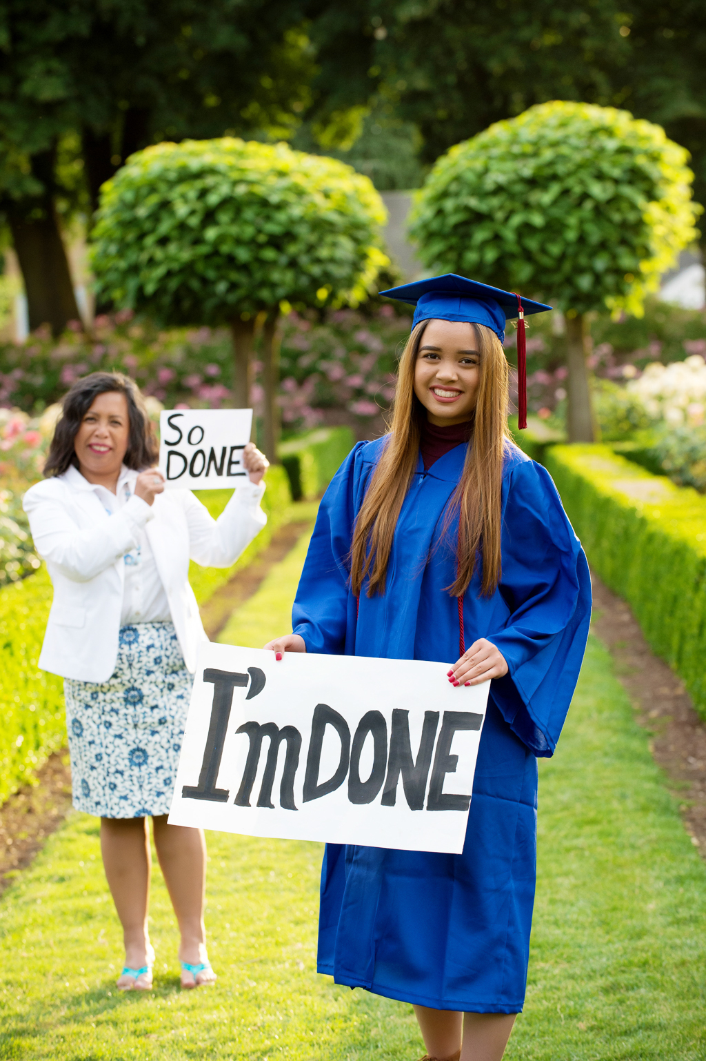 a high school senior wears her cap and gown and holds a sign that says I'm Done and her mom in the background holds a sign that says So Done
